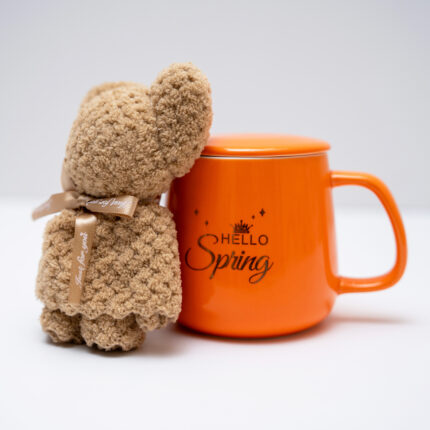 Bright orange mug with 'Hello Spring' print next to a plush teddy bear with a bow on a white background.