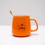 Bright orange mug with 'Hello Spring' print next to a plush teddy bear with a bow on a white background.