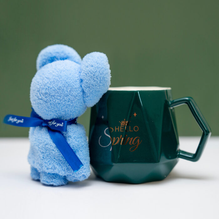 Dark green mug with 'Hello Spring' print beside a blue teddy bear with a blue ribbon on a green background.