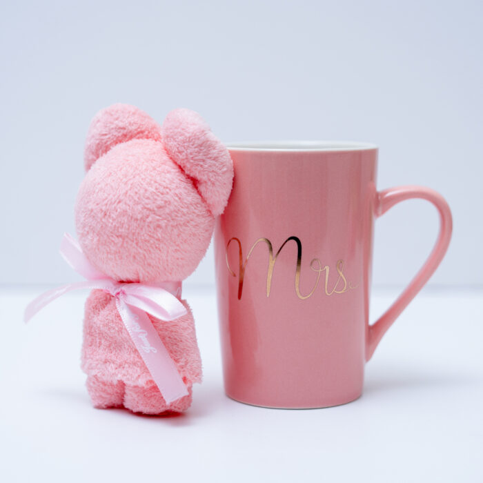 A pink teddy bear standing next to a pink mug with "Mrs" written in gold cursive