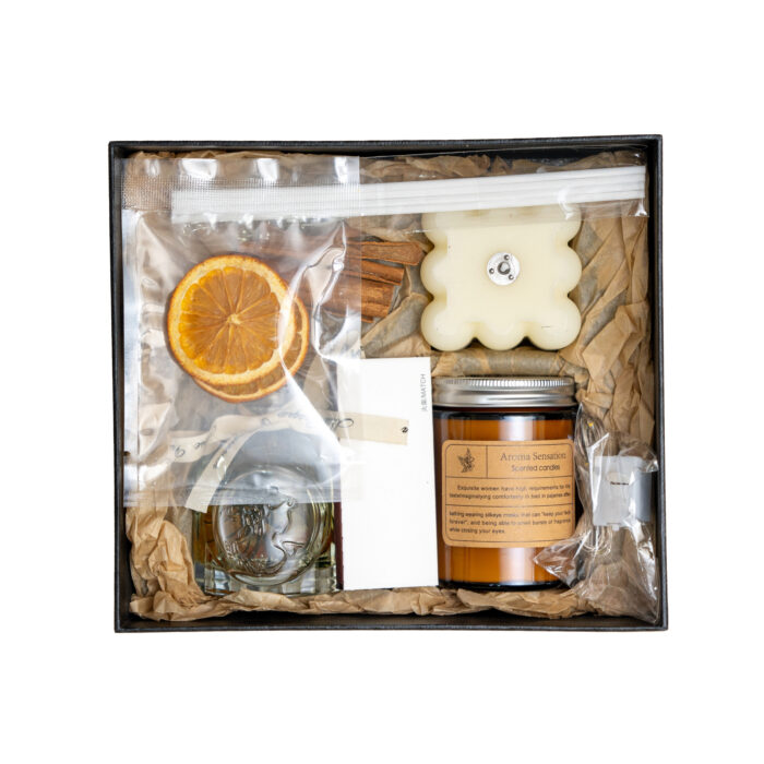 Gift set with aromatherapy products including a candle, bar soap, glass perfume bottle, and dried orange slice, packaged in a clear-topped box with crinkled paper