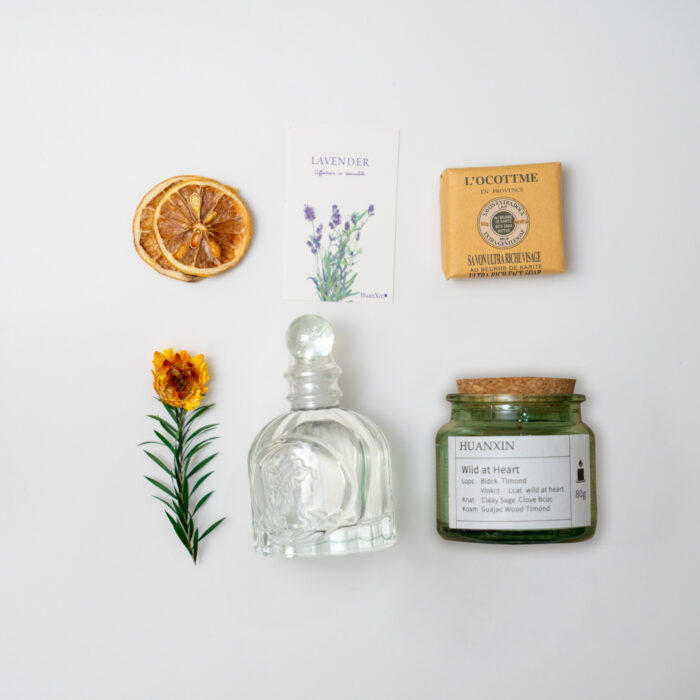 "Flat lay of aromatherapy items including dried orange slice, lavender card, bar soap, glass perfume bottle, and a jar candle on a burlap background